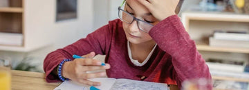 Child in red shirt nervoously looking at notebook while holding a blue pen
