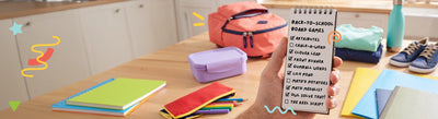 A hand holding a notepad with a SimplyFun back-to-school games checklist, surrounded by colorful school supplies and a pink backpack on a wooden table in a kitchen setting.