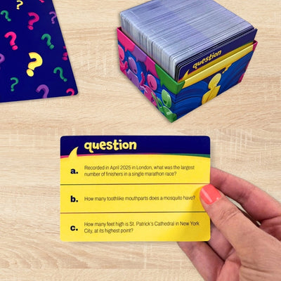 A hand with pink nail polish holds a yellow trivia question card in front of a colorful card box and a stack of cards on a light wood tabletop.