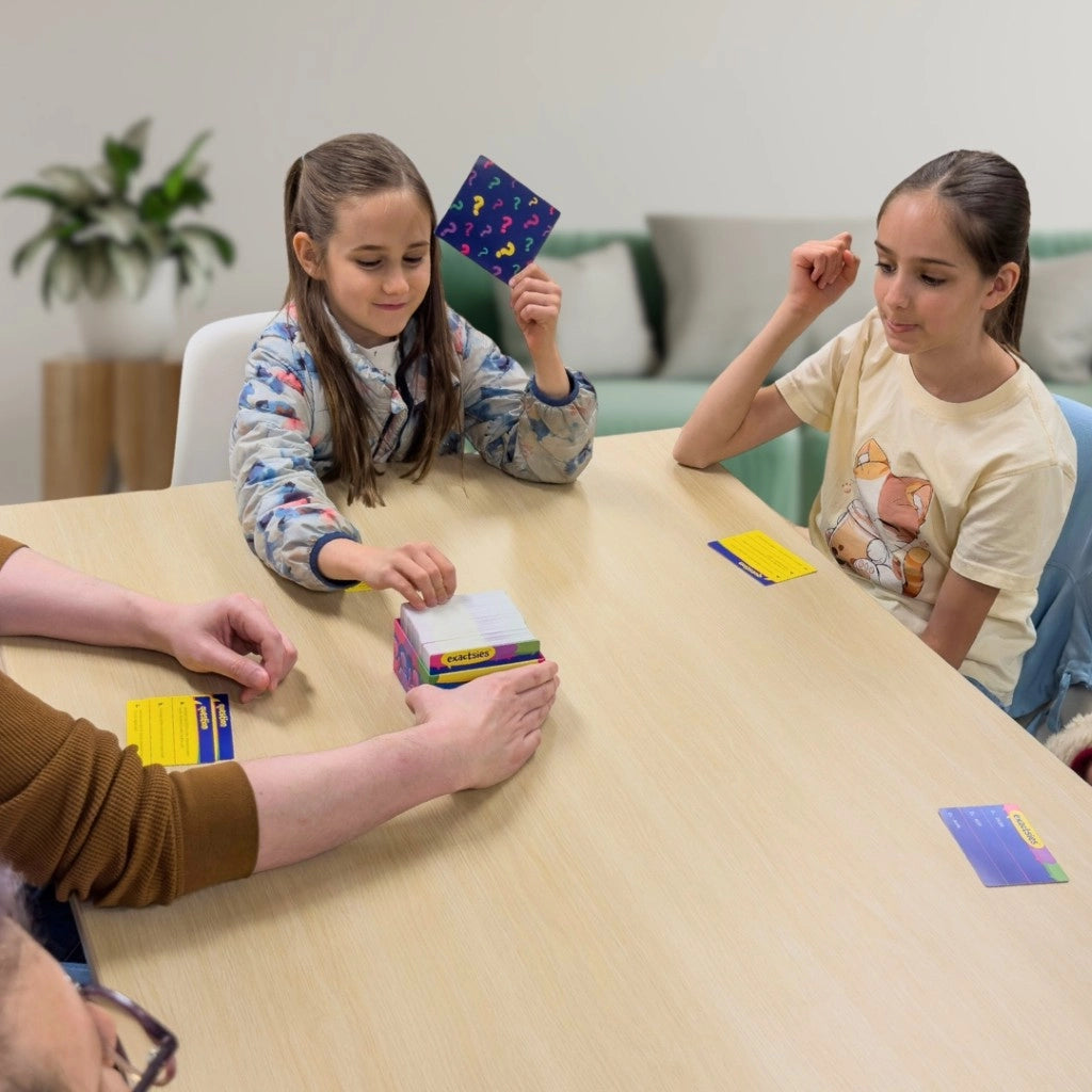 Three people seated around a light wood table in a bright indoor setting, interacting with colorful card games or cards. A child holds up a purple card, another child sits to the right, and hands at the table are interacting with a stack of cards.-SimplyFun