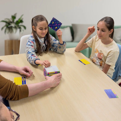 Three people seated around a light wood table in a bright indoor setting, interacting with colorful card games or cards. A child holds up a purple card, another child sits to the right, and hands at the table are interacting with a stack of cards.